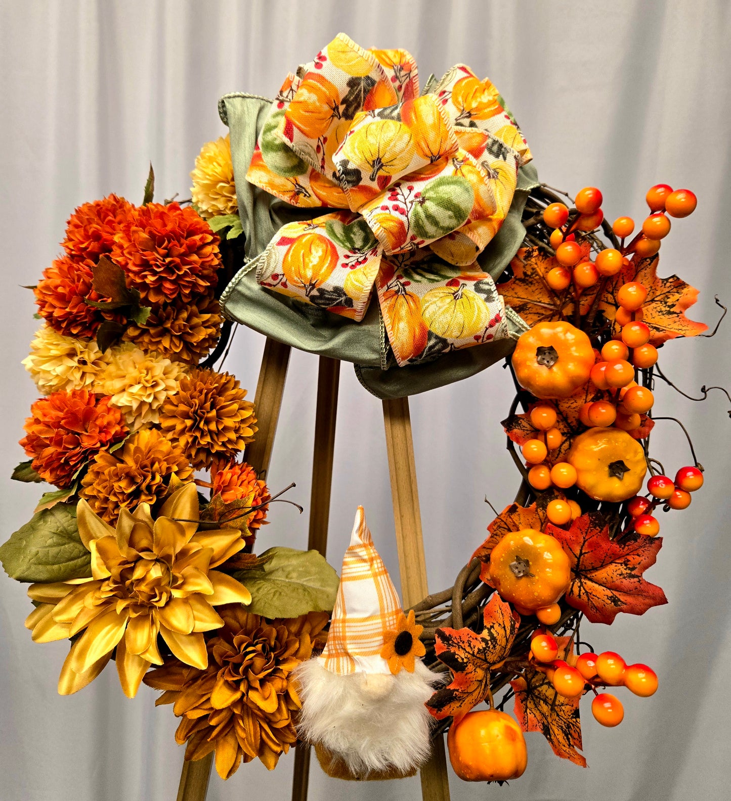 12″ grapevine wreath featuring autumn florals in yellow, orange, and brown, a gnome accent, pumpkin berry picks, and a multi-ribbon bow