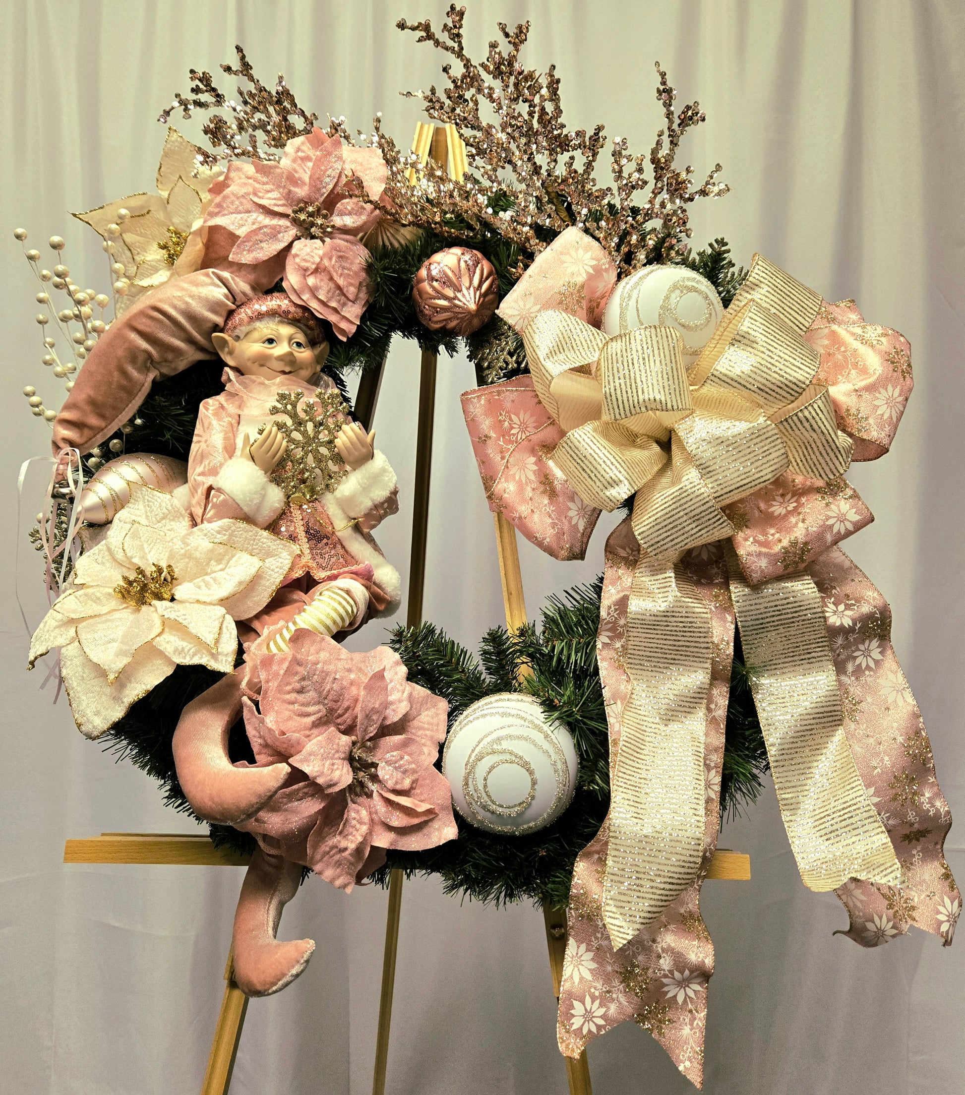 22″ Christmas wreath in pink, cream & gold featuring an elf figure, poinsettias, ornament balls, gold picks, and a multi-ribbon bow
