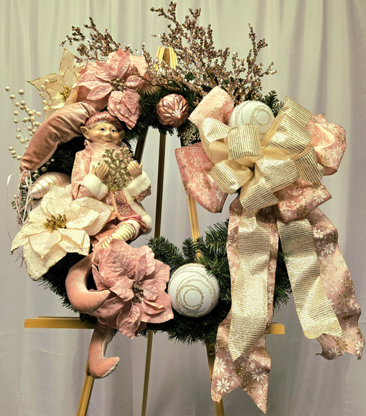 22″ Christmas wreath in pink, cream & gold featuring an elf figure, poinsettias, ornament balls, gold picks, and a multi-ribbon bow
